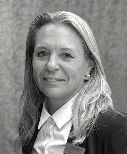 A woman with long hair smiles, wearing a suit and a name tag against a textured background.